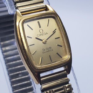 May include: A gold-tone Omega De Ville quartz wristwatch with a rectangular case and a matching gold-tone expansion link bracelet. The watch face features a gold dial with thin black hour markers, black hands, and the text Omega, De Ville, Quartz, Swiss Made.