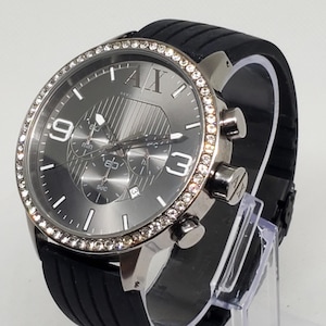 May include: A silver-toned wristwatch with a black rubber band. The watch face is gray with silver hands and markers, and a bezel encrusted with small, clear crystals. The watch has three sub-dials and the letters "AX" at the top.