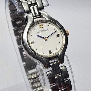 Puede incluir: Un reloj de pulsera Courr&egrave;ges plateado con esfera blanca, detalles dorados y una correa de eslabones plateados. La esfera del reloj muestra el logotipo de Courr&egrave;ges y agujas azules. El reloj se exhibe sobre un soporte de pl&aacute;stico transparente.