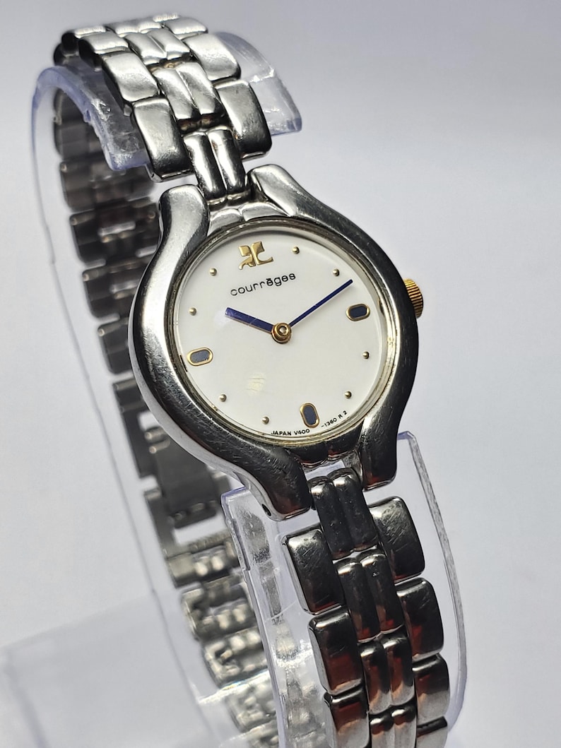 Puede incluir: Un reloj de pulsera Courr&egrave;ges plateado con esfera blanca, detalles dorados y una correa de eslabones plateados. La esfera del reloj muestra el logotipo de Courr&egrave;ges y agujas azules. El reloj se exhibe sobre un soporte de pl&aacute;stico transparente.