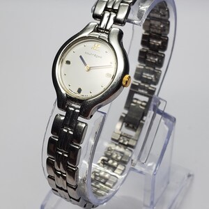 Puede incluir: Un reloj de pulsera Courr&egrave;ges con una correa de metal plateado y una esfera redonda. La esfera del reloj es plateada con detalles dorados y el logotipo de Courr&egrave;ges. El reloj se exhibe sobre un soporte de pl&aacute;stico transparente.