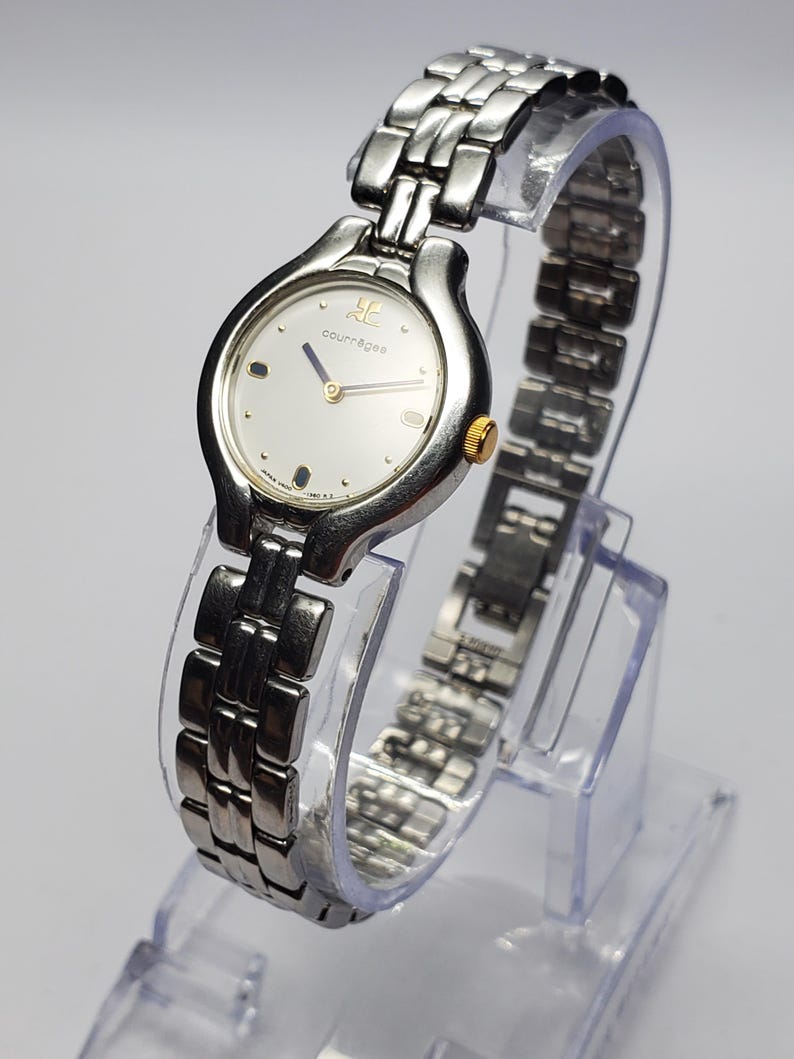 Puede incluir: Un reloj de pulsera Courr&egrave;ges con una correa de metal plateado y una esfera redonda. La esfera del reloj es plateada con detalles dorados y el logotipo de Courr&egrave;ges. El reloj se exhibe sobre un soporte de pl&aacute;stico transparente.