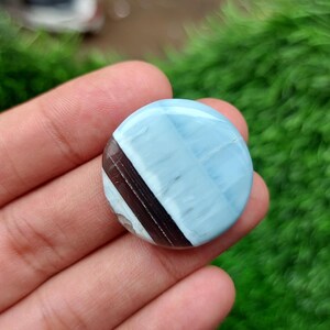May include: A round, polished gemstone with a light blue and brown color scheme. The stone features a diagonal band of dark brown across a light blue background. The gemstone is held in a hand, with a green background.
