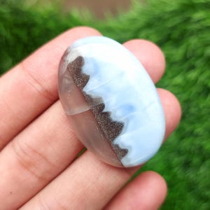 May include: Oval-shaped gemstone with a polished surface. The stone features a blend of light blue, brown, and white colors. The gemstone is held in a hand against a blurred green background.