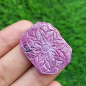 May include: A carved, pink gemstone with a floral design. The gemstone is a rectangular shape with beveled edges. The floral design is carved into the surface of the stone, with detailed petals radiating from the center.