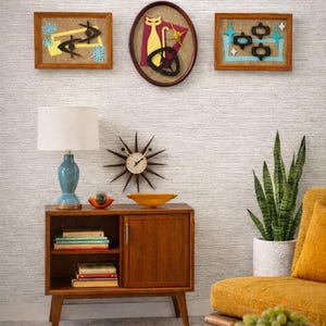 Può includere: Una scena di soggiorno moderno di metà secolo con opere d'arte incorniciate, un mobile in legno con una lampada blu e un orologio a stella. Una poltrona gialla e un tavolino con una ciotola di uva completano l'arredamento retrò.