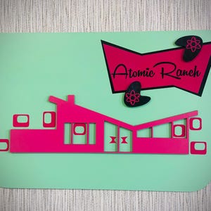Puede incluir: Un letrero de estilo retro con un fondo verde menta y una silueta de casa moderna de mediados de siglo en rosa intenso. El letrero dice "Atomic Ranch" en escritura negra, con formas atómicas. Una pieza única de decoración del hogar.