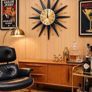 May include: A starburst wall clock featuring a circular gold face with black numerals and radiating dark blue pointed spikes tipped with small gold spheres. The clock hands are black and positioned at the twelve o'clock mark on the dial.