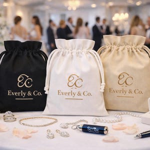 May include: Three drawstring bags in black, white, and beige, each with the gold text "Everly & Co." displayed. Various jewelry items, including rings, bracelets, and earrings, are arranged in front of the bags. A pen and a small black object are also visible.