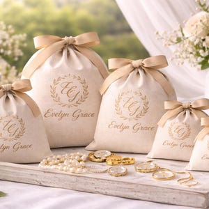 May include: Cream-colored drawstring bags with gold bows, featuring a gold wreath and the text "Evelyn Grace." The bags are arranged on a wooden tray with jewelry, including necklaces and rings. A candle and flowers are in the background.