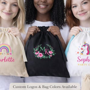 May include: Three drawstring bags in beige, black, and white. The beige bag has a rainbow graphic and the name "Charlotte". The black bag features a floral wreath with the initials "MK". The white bag displays a unicorn and the name "Sophia".
