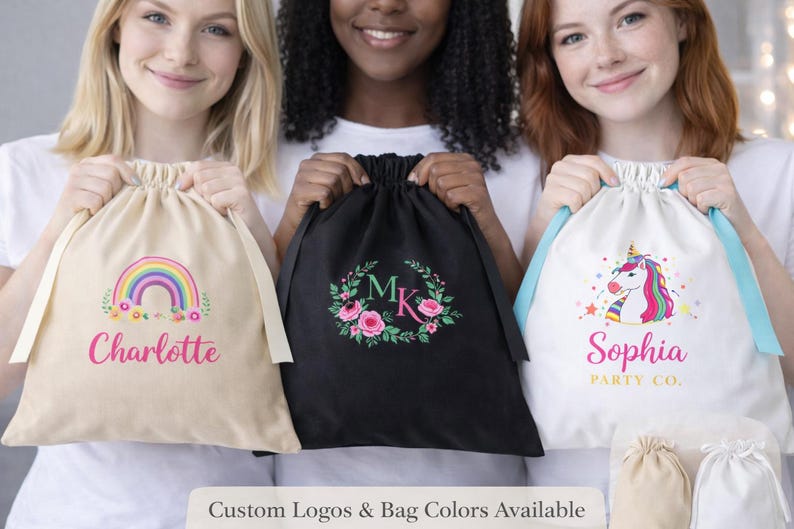 May include: Three drawstring bags in beige, black, and white. The beige bag has a rainbow graphic and the name "Charlotte". The black bag features a floral wreath with the initials "MK". The white bag displays a unicorn and the name "Sophia".