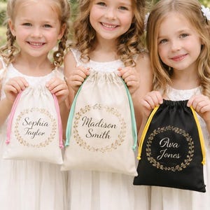 May include: Three young girls holding personalized drawstring bags. The bags are in cream and black, with gold text and floral wreath designs. The bags have drawstrings in pink, mint green, and yellow. The names Sophia Taylor, Madison Smith, and Ava Jones are printed on the bags.