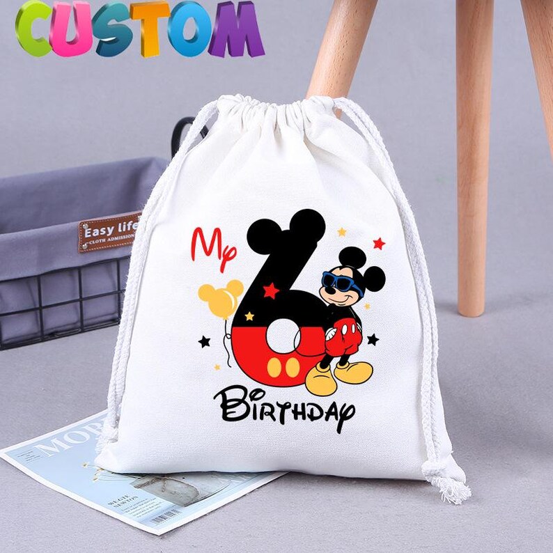 May include: White drawstring bag with a Mickey Mouse design. The bag has a red and black number six with Mickey Mouse wearing sunglasses and a red and black outfit. The text "My 6th Birthday" is printed on the bag.