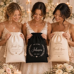 May include: Three drawstring gift bags made of fabric. The left bag is cream with gold text reading Thank You For Celebrating Our Special Day. The center bag is black with gold text reading Mr. & Mrs. Johnson Est. 2025. The right bag is cream with gold text reading With Love Thank You For Sharing Our Wedding Day.