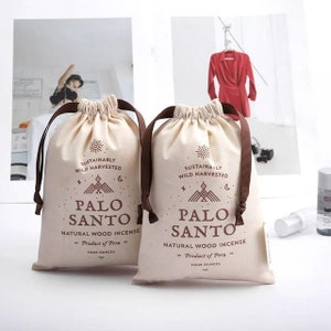 May include: Two white drawstring bags with brown drawstrings. Each bag has a brown logo that says "Sustainably Wild Harvested" and "Palo Santo Natural Wood Incense" with a sun, moon, and mountain design. The text "Product of Peru" and "Four Ounces" are also printed on the bags.