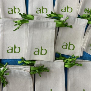 May include: A collection of off-white canvas drawstring bags, each printed with the green letters "ab". The bags are tied with green ribbon. The bags are arranged on a blue surface.
