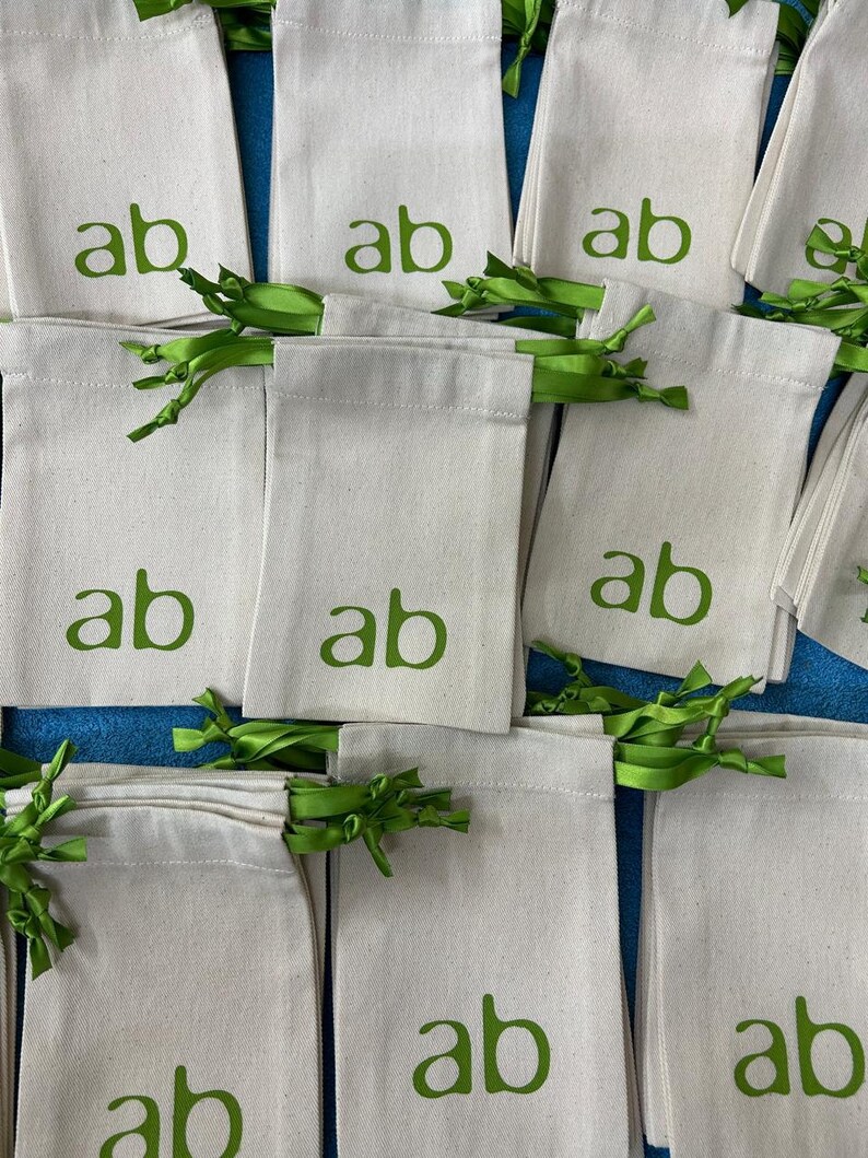 May include: A collection of off-white canvas drawstring bags, each printed with the green letters "ab". The bags are tied with green ribbon. The bags are arranged on a blue surface.