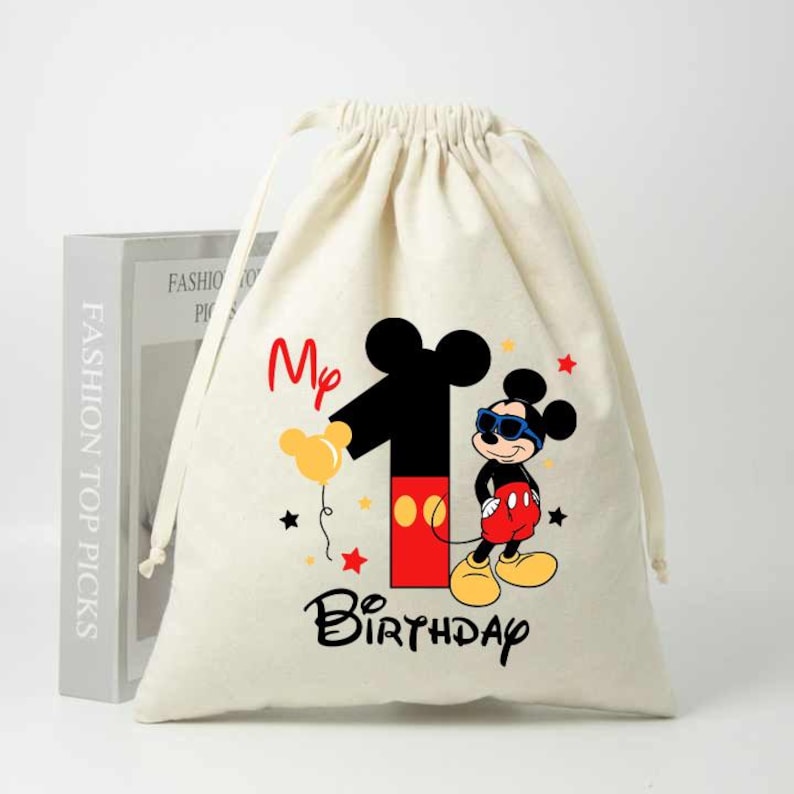 May include: A white drawstring bag with a Mickey Mouse design. The bag features a red and black number one with Mickey Mouse wearing sunglasses and holding a yellow balloon. The text "My 1st Birthday" is printed below the design.