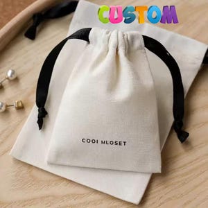 Puede incluir: Dos bolsas de algodón blanco con cordones de ajuste negros. La bolsa más pequeña tiene el texto "CUSTOM" impreso en colores arcoíris y la bolsa más grande tiene el texto "COOOI MLOSET" impreso en negro.