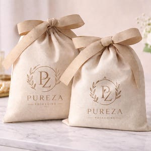 May include: Two beige drawstring bags with tan ribbon bows. Each bag has a gold logo that reads "PZ PUREZA PACKAGING". The bags are on a marble surface, with a vase of white roses in the background.