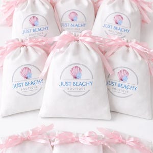 Op de afbeelding: Witte trekkoordzakken met roze linten. Elke tas heeft een cirkelvormig logo met een zeeschelp en de tekst "JUST BEACHY BOUTIQUE". De tassen zijn gegroepeerd, mogelijk voor een productpresentatie.