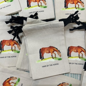 Op de afbeelding: Meerdere kleine witte canvas tassen met zwarte trekkoorden. Elke tas heeft een aquarel illustratie van een bruin paard met de tekst "Hair of the Horse" eronder gedrukt.