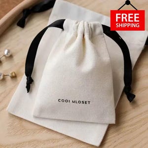 Puede incluir: Dos bolsas con cordón de color blanquecino con cierres de cinta negra. La bolsa más pequeña está encima de una bolsa rectangular más grande. El texto "COOI MLOSET" está impreso en la parte delantera de la bolsa más pequeña. Un letrero rojo con las palabras "FREE SHIPPING" es visible.