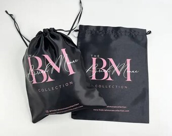 May include: Two black satin drawstring bags with pink text that reads "The Briah Monae Collection." One bag is closed, and the other is open. The bags have a shiny finish and are likely used for packaging or storage.
