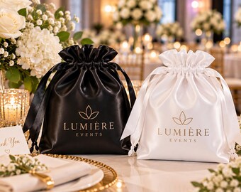 May include: Two drawstring pouches, one black and one white, made of a smooth, satin-like fabric. Each pouch features a gold-colored logo with a stylized leaf icon above the text "LUMIÈRE EVENTS" printed clearly on the front center.