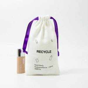 Puede incluir: Bolsa de algodón blanco con cordón de ajuste y cintas de color morado. La bolsa tiene la palabra "RECYCLE" impresa en la parte delantera, junto con pequeñas ilustraciones de botellas y un pez. La bolsa también tiene el texto "#smartbeauty #footwastecollection #美丽新主张" impreso en la parte delantera.