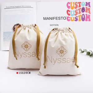 May include: Two white cotton drawstring bags with gold ribbon ties and a gold logo that says "Nyssae" with a decorative floral design. The bags are 15x20 cm.