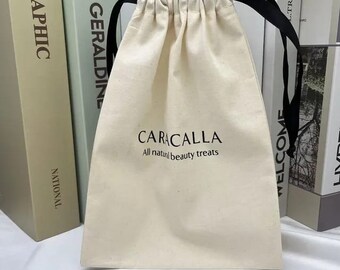 Może przedstawiać: Biały bawełniany worek ze sznurkiem do ściągania w kolorze czarnym. Na przodzie worka znajduje się napis "CARACALLA All natural beauty treats".