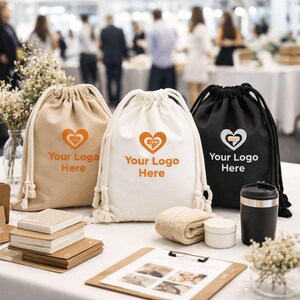 May include: Three drawstring bags in beige, white, and black, each with a heart-shaped logo and the text "Your Logo Here" in orange or white. The bags are on a white table with other items, including a towel, cream jar, and a black travel mug.