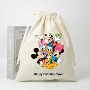 May include: A white cotton drawstring bag with a colorful print of Mickey Mouse, Minnie Mouse, Donald Duck, Goofy, Pluto, and Daisy Duck. The bag has the text "Happy Birthday Ryan!" printed on the front.
