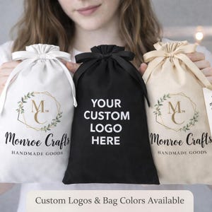 May include: Three drawstring bags in white, black, and beige. The white and beige bags feature a logo with the text "Monroe Crafts HANDMADE GOODS". The black bag has the text "YOUR CUSTOM LOGO HERE". The text "Custom Logos & Bag Colors Available" is also present.