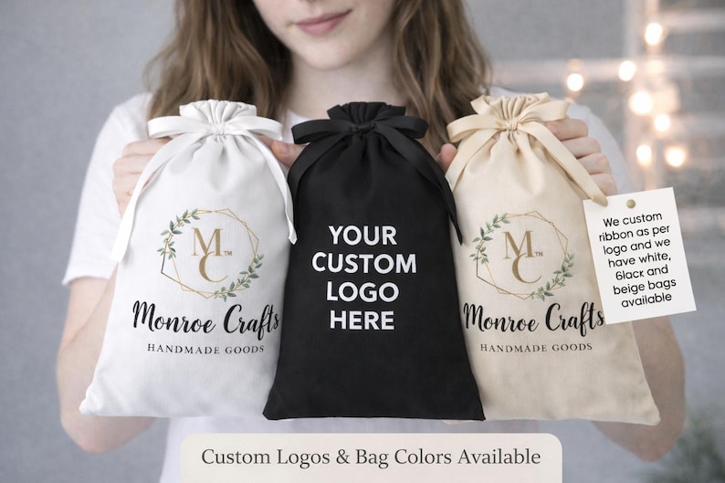 May include: Three drawstring bags in white, black, and beige. The white and beige bags feature a logo with the text "Monroe Crafts HANDMADE GOODS". The black bag has the text "YOUR CUSTOM LOGO HERE". The text "Custom Logos & Bag Colors Available" is also present.