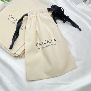 Puede incluir: Una pila de bolsas de algodón blanco con cordón y lazos de cinta negra. Las bolsas tienen el texto "CAIACALLA All natural beauty treats" impreso en ellas.