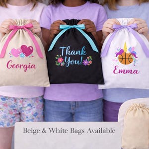 May include: Three drawstring bags in beige, black, and white. The beige bag has pink hearts and the name "Georgia". The black bag says "Thank You!" with floral accents. The white bag features a unicorn, basketball, and the name "Emma".