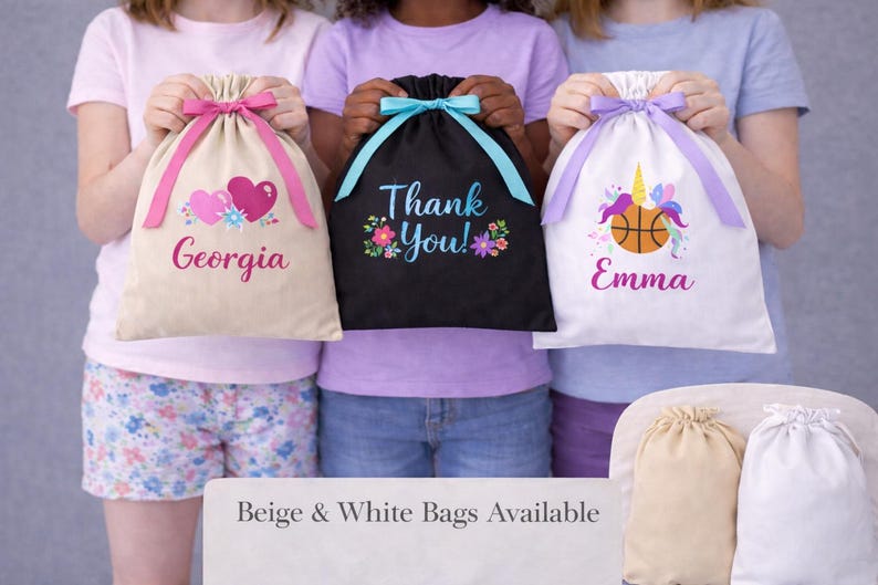 May include: Three drawstring bags in beige, black, and white. The beige bag has pink hearts and the name "Georgia". The black bag says "Thank You!" with floral accents. The white bag features a unicorn, basketball, and the name "Emma".