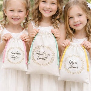 May include: Three young girls in white dresses hold personalized drawstring bags. Each bag has a name in elegant script within a gold wreath. The bags have pink, mint green, and yellow drawstrings.