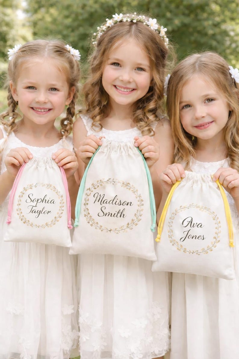 May include: Three young girls in white dresses hold personalized drawstring bags. Each bag has a name in elegant script within a gold wreath. The bags have pink, mint green, and yellow drawstrings.