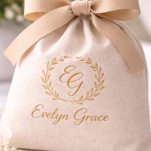 May include: A beige drawstring bag with a matching ribbon bow at the top. The bag features a gold-colored wreath design with the initials "E G" and the name "Evelyn Grace" printed below. The bag is made of a textured fabric.