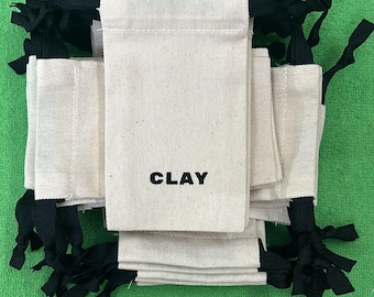 May include: A stack of 10 small, off-white canvas bags with black drawstrings. The bags have the word "CLAY" printed in black on the front.