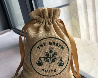 Op de afbeelding: Een bruine jute trekkoordtas met een gouden lint. De tas heeft een groen cirkellogo met de tekst "The Green Suite" met een gestileerd planten- en weegschaalontwerp.
