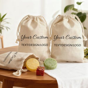 May include: Two beige drawstring bags with the text "Your Custom TEXT/DESIGN/LOGO". A smaller bag is open, revealing a yellow and a green soap, and a small red gift box. The soaps have floral designs.