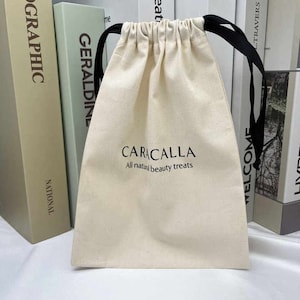 Puede incluir: Una bolsa de algodón blanco con cordones de ajuste negros. La bolsa tiene el texto "CARACALLA All natural beauty treats" impreso en la parte delantera.