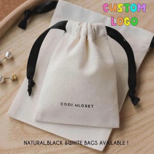 May include: A natural-colored canvas drawstring pouch featuring black ribbon ties and the text "COOIMLOSET" printed in black on the front. The pouch is positioned on a larger, matching flat canvas bag, both made of a woven fabric material.