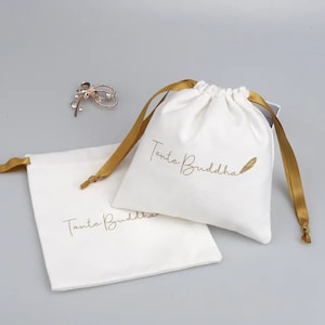 Puede incluir: Dos bolsas de algodón blanco con cordón de ajuste y cintas doradas. Las bolsas tienen el texto "Tante Buddha" impreso en dorado en la parte delantera. Una bolsa tiene un diseño de pluma dorada junto al texto.