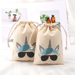 Puede incluir: Dos pequeñas bolsas de cordón beige con un diseño de unicornio. El unicornio tiene una melena azul y lleva gafas de sol. Las bolsas están hechas de algodón y tienen un cierre de cordón.